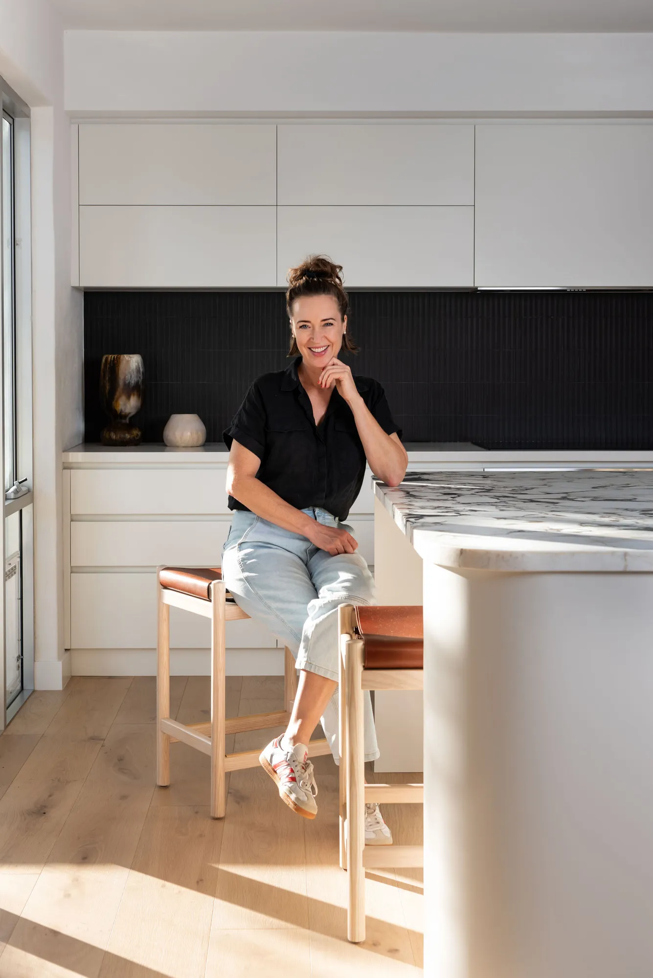 Lisa Ewart of Collected Interiors seated at a marble island bench in a Claremont Perth kitchen with white cabinetry and a black fluted tile splashback.